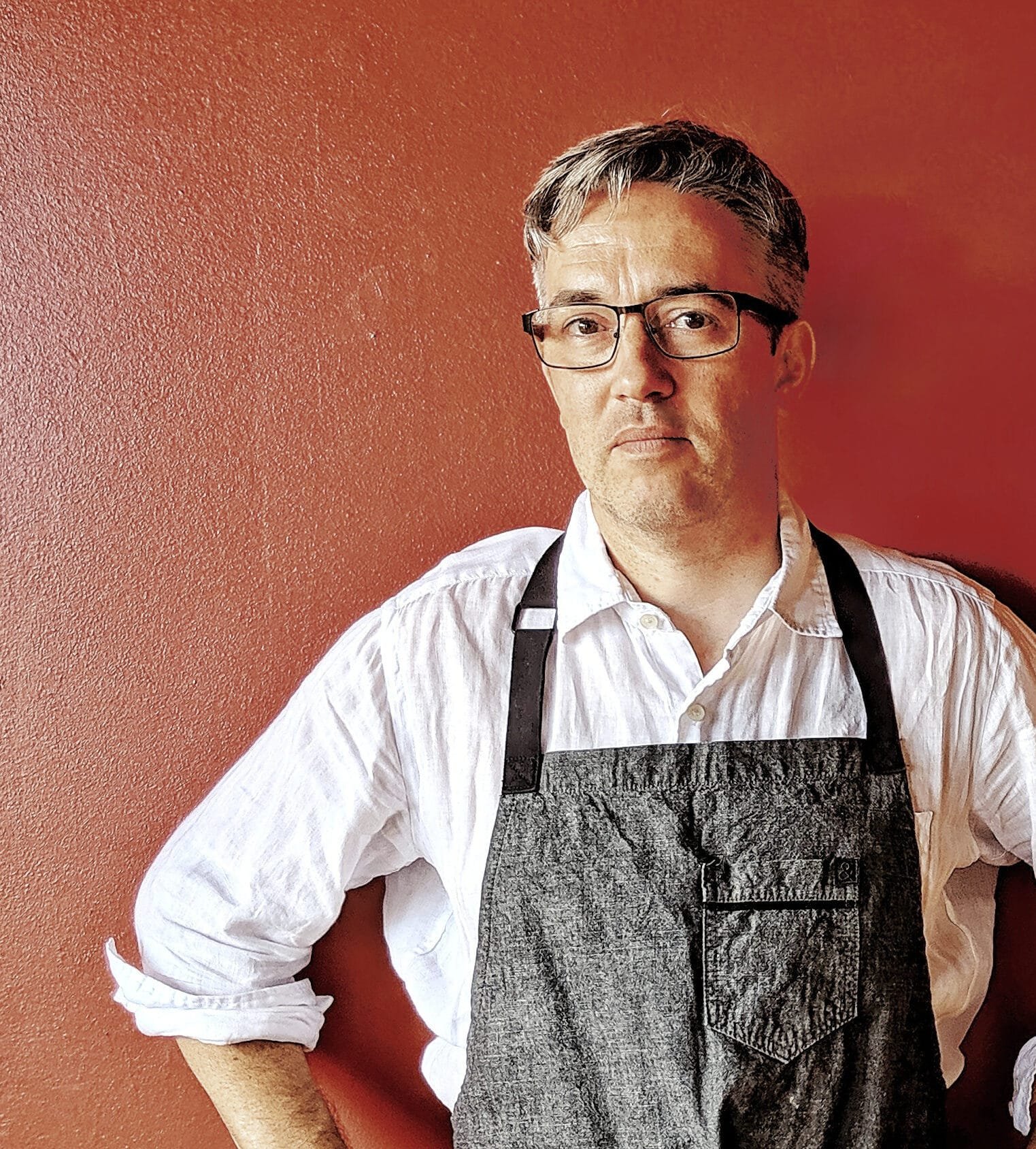 Medium portrait photograph of Eiten, the creator of Tenzo's Kitchen, with gray-streaked hair and glasses, wearing a white button-down shirt and a denim-style apron. He stands with hands on hips and a serious expression against a textured red wall.